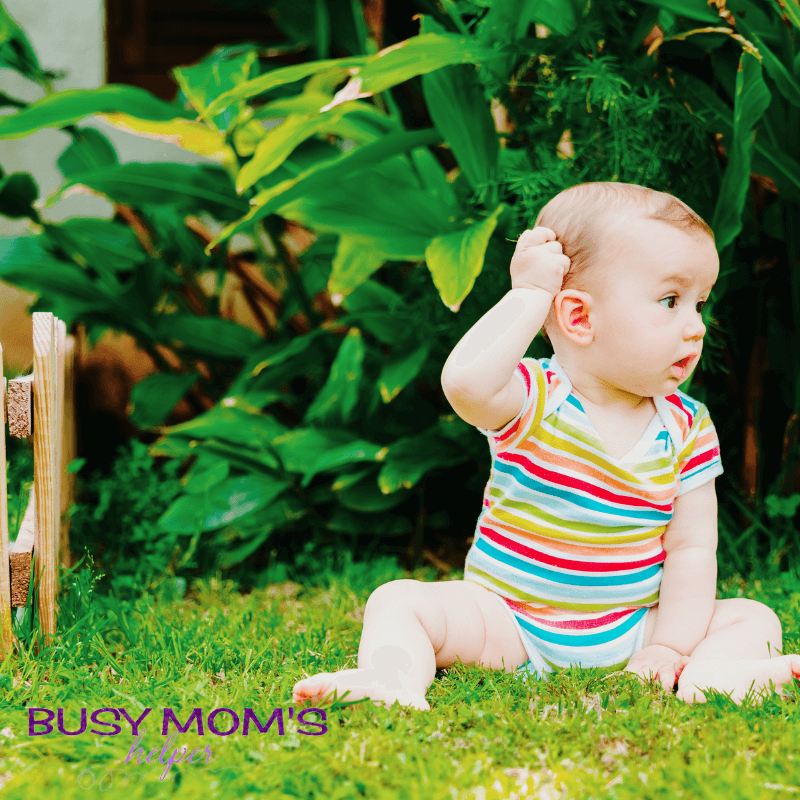 photo of a baby scratching head while sitting on grass outside.